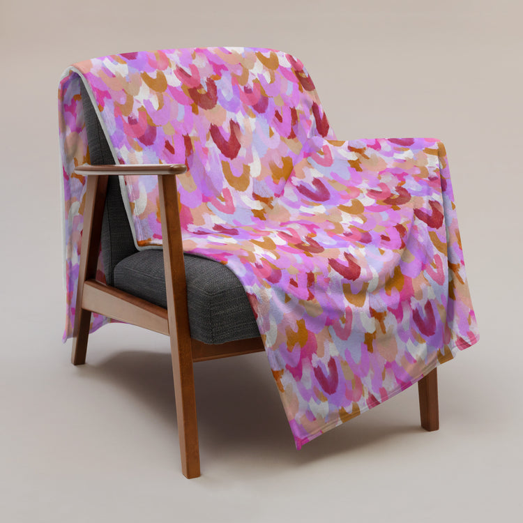 A wooden chair with a gray cushion is draped with My Favourite Colour is Rainbows Pink Rainbow Throw Blanket, featuring pink, purple, and orange abstract patterns. This cozy blanket enhances the stylish décor against the plain, neutral background.