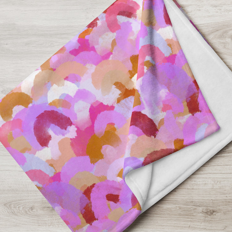 A folded Pink Rainbow Throw Blanket by My Favourite Colour is Rainbow, featuring a colorful abstract pattern of pink, purple, orange, and white brushstrokes, stands out stylishly against a light wooden surface.