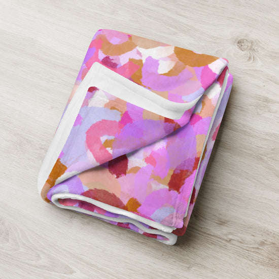 The Pink Rainbow Throw Blanket by My Favourite Colour is Rainbow features a vibrant abstract pattern with overlapping watercolor-style circles in shades of pink, purple, orange, and beige on a light wood surface, making it a stylish décor piece for any room.