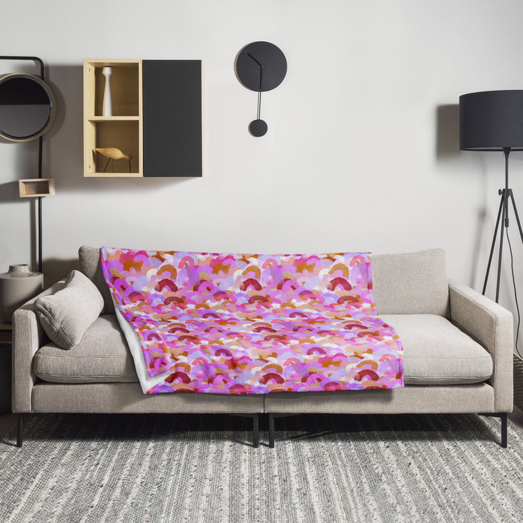 A modern living room is elevated by My Favourite Colour is Rainbows Pink Rainbow Throw Blanket, featuring a pink, red, and orange abstract pattern. The décor includes a minimalist clock, black floor lamp, and decorative shelf items. A gray rug adds texture to the beige sofa area.