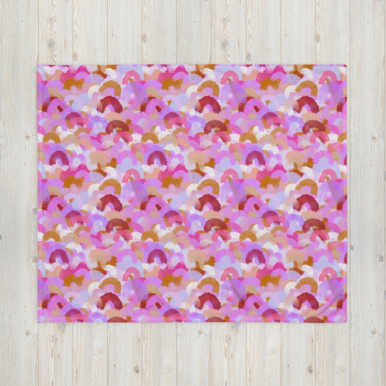 The Pink Rainbow Throw Blanket by My Favourite Colour is Rainbow showcases a vibrant pattern of abstract semicircles in pink, orange, red, and white. Its cozy texture enhances any light wooden surface with visible grain, making it perfect for stylish décor.