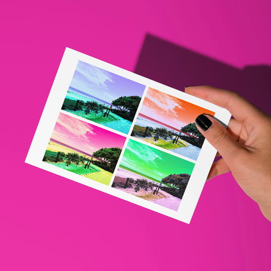 A hand with black nail polish holds a Pohutukawa Pop Art greeting card (Version 2) from My Store, featuring four colorful beach scenes. A vivid pink background adds depth and vibrancy to the New Zealand-inspired design.