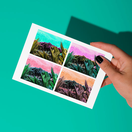 A hand with black nail polish holds the My Stores Pohutukawa Pop Art greeting card (Version 3) showcasing four tropical scenes with vibrant turquoise, pink, purple, and orange skies on a teal background, inspired by New Zealands stunning vistas.
