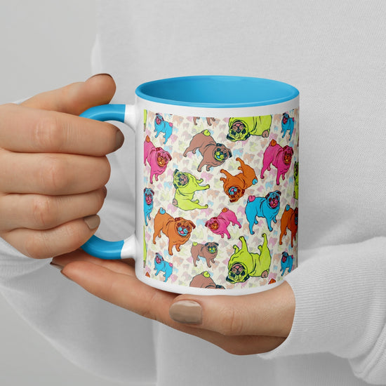 A person wearing a white sweatshirt holds a Pugalicious Mug by My Favourite Colour is Rainbow, featuring vibrant cartoon pugs in green, blue, pink, and brown and a light blue interior and handle—perfect for pug enthusiasts.