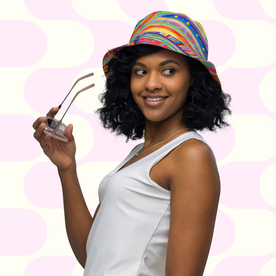 A woman with shoulder-length curly hair wears The Rainbow Connection/Summer Sorbet Reversible Bucket Hat by My Favourite Colour is Rainbow. In a white sleeveless top, she holds glasses, smiling and gazing sideways against a pink and white geometric pattern, flaunting her style BFF.