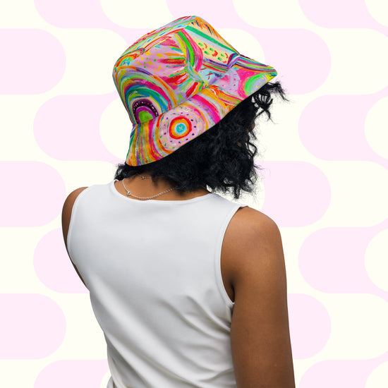 Someone with curly hair is seen wearing The Rainbow Connection/Summer Sorbet Reversible Bucket Hat by My Favourite Colour is Rainbow and a white sleeveless top. Theyre facing away from the camera against a pink and white geometric background, exuding an effortless BFF style vibe.