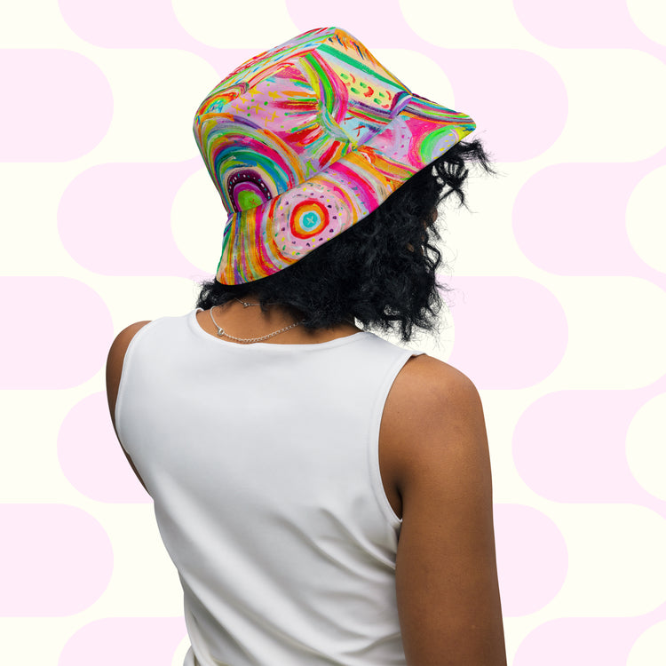Someone with curly hair is seen wearing The Rainbow Connection/Summer Sorbet Reversible Bucket Hat by My Favourite Colour is Rainbow and a white sleeveless top. Theyre facing away from the camera against a pink and white geometric background, exuding an effortless BFF style vibe.
