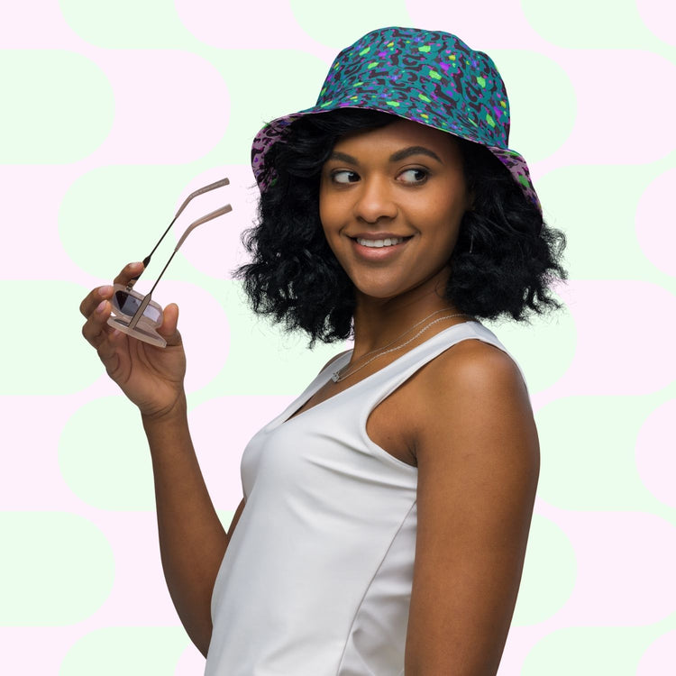A woman in a white tank top holds sunglasses and smiles sideways, stylishly wearing the My Favourite Colour is Rainbows Silly Sausages Green/Pink Reversible Bucket Hat against a pastel geometric background.