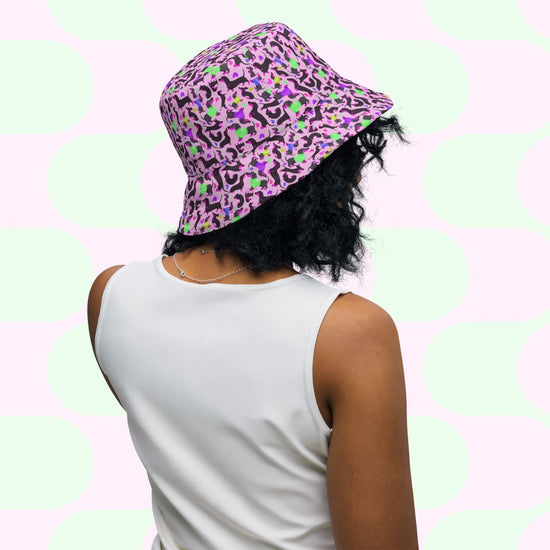 A person faces away, wearing a white sleeveless top and the Silly Sausages Green/Pink Reversible Bucket Hat by My Favourite Colour is Rainbow. The backdrop features large pastel green and pink circles, enhancing the stylish and comfortable vibe.