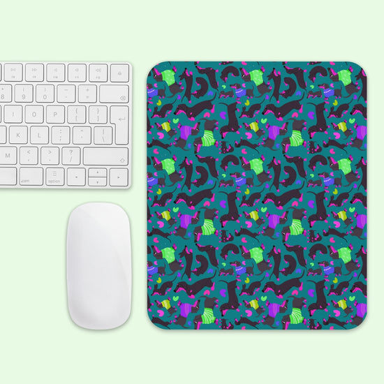 A white computer keyboard and mouse on a light green surface with the Silly Sausages Mouse Pad by My Favourite Colour is Rainbow, featuring abstract shapes and vibrant dinosaurs in purple, green, and blue.