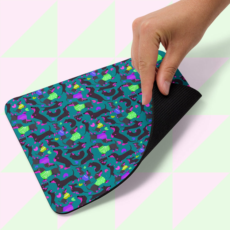 A hand holds the Silly Sausages Mouse Pad by My Favourite Colour is Rainbow, featuring dinosaurs in shades of blue, green, and purple on a dark background with alternating pastel pink and green triangles.
