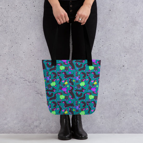 A person carries a vibrant My Favourite Colour is Rainbow Silly Sausages Tote Bag, with a fun pattern of black-and-pink dachshunds wearing green and purple jumpers on a dark teal background. They're wearing black pants and boots, standing against a textured gray wall.