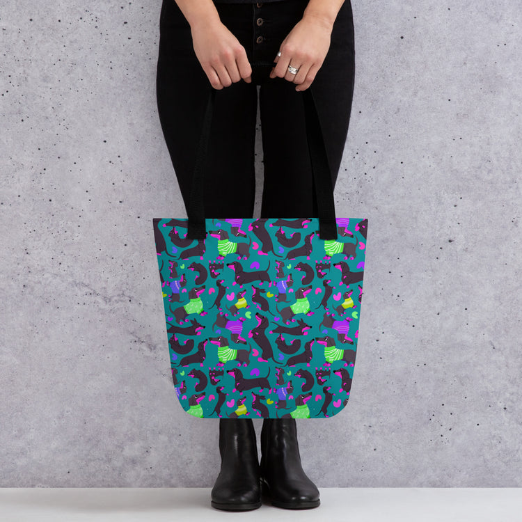 A person carries a vibrant My Favourite Colour is Rainbow Silly Sausages Tote Bag, with a fun pattern of black-and-pink dachshunds wearing green and purple jumpers on a dark teal background. They're wearing black pants and boots, standing against a textured gray wall.