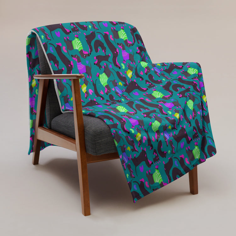 A wooden armchair with a dark gray cushion is covered by the My Favourite Colour is Rainbows Silly Sausages Throw Blanket, featuring playful green, purple, and teal dinosaurs on a dark background. A pink throw adds charm while both are set against a light gray backdrop.