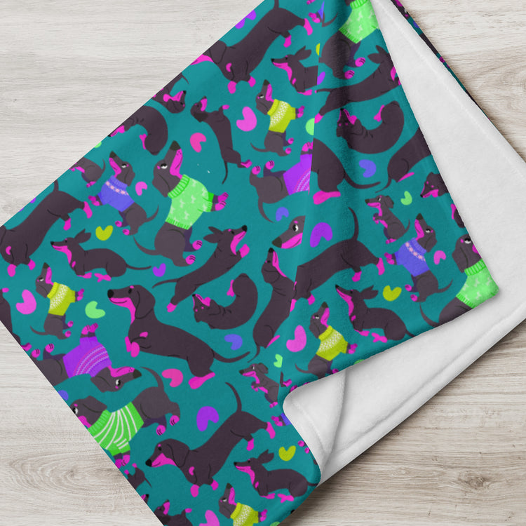 The Silly Sausages Throw Blanket by My Favourite Colour is Rainbow showcases playful dachshunds in colorful sweaters against a teal background with pink and purple hearts. The cozy reverse is pure white, perfect for a cheerful room.
