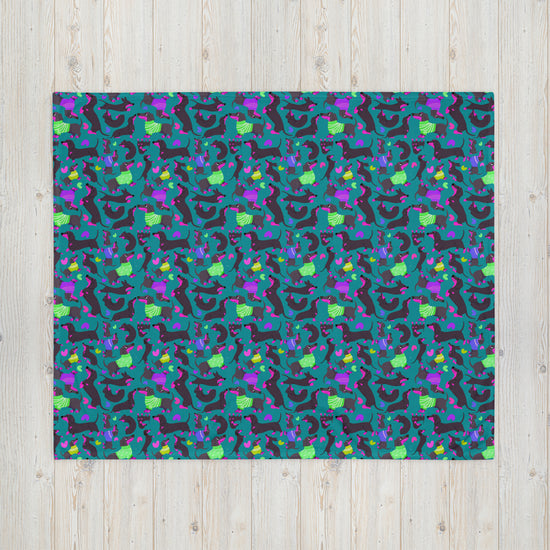 The dark background features an abstract pattern with purple, green, yellow, and blue geometric shapes. Beside it lies the cozy Silly Sausages Throw Blanket in green from My Favourite Colour is Rainbow. The setup rests on a light wooden surface, enhancing the vibrant visual effect.