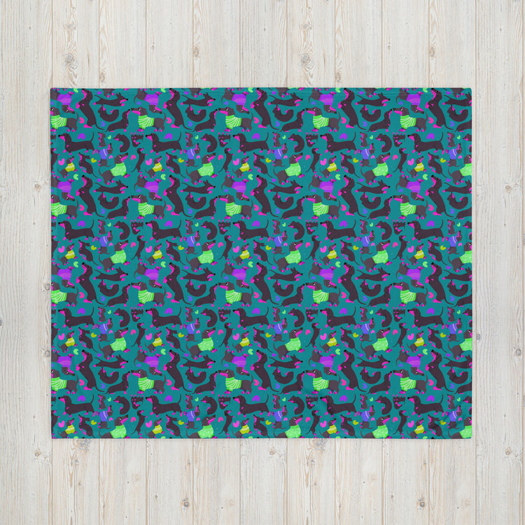 The dark background features an abstract pattern with purple, green, yellow, and blue geometric shapes. Beside it lies the cozy Silly Sausages Throw Blanket in green from My Favourite Colour is Rainbow. The setup rests on a light wooden surface, enhancing the vibrant visual effect.