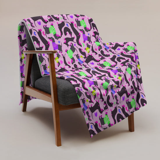 The wooden armchair with a dark gray cushion is enhanced by the Silly Sausages Throw Blanket - Pink from My Favourite Colour is Rainbow, adorned with a playful pattern in pink, purple, green, and black flamingoes. The backdrop is a neutral light gray.