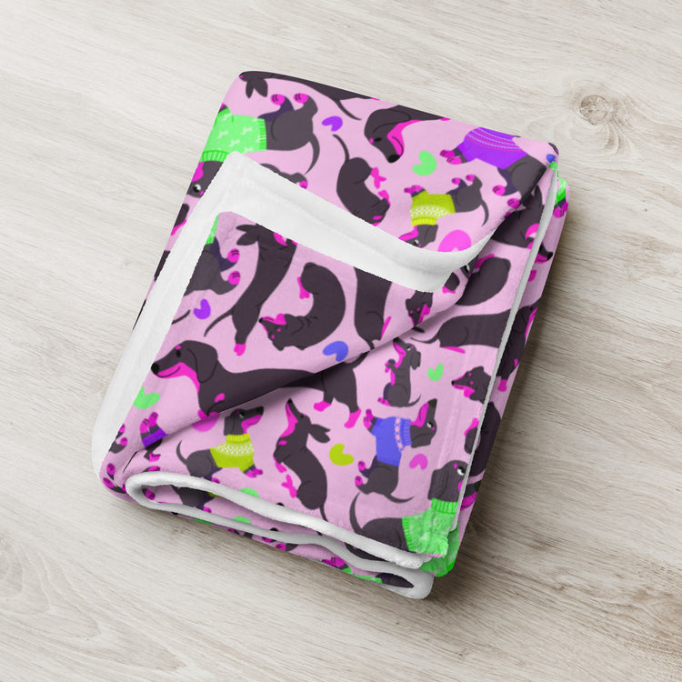 The Silly Sausages Throw Blanket - Pink by My Favourite Colour is Rainbow showcases black dachshunds in colorful sweaters on a pink backdrop, playfully arranged as Silly Sausages, resting on a light wood surface.