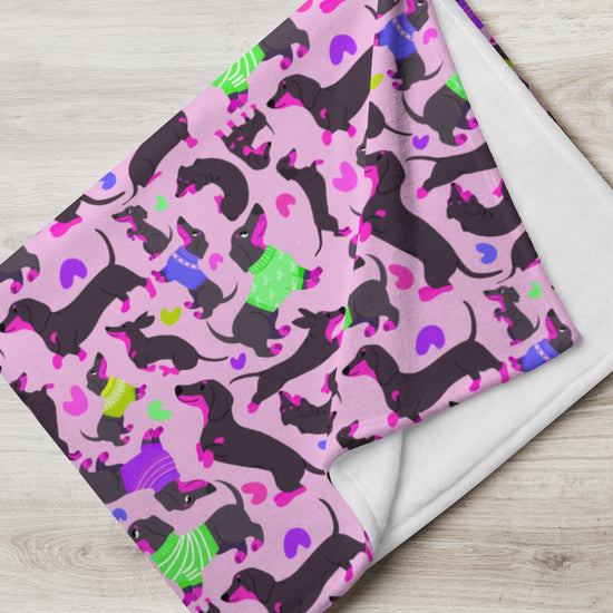 Discover the playful Silly Sausages Throw Blanket - Pink by My Favourite Colour is Rainbow, showcasing black dachshunds in colorful sweaters, hats, and boots on a pink backdrop with purple, green, and blue hearts—a charming addition to any room!.