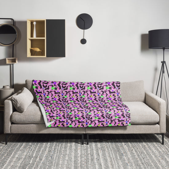 A modern living room features a light gray sofa with the whimsical Silly Sausages Throw Blanket in pink by My Favourite Colour is Rainbow. The background showcases a black floor lamp, decorative shelves, and a round mirror on the light gray wall.