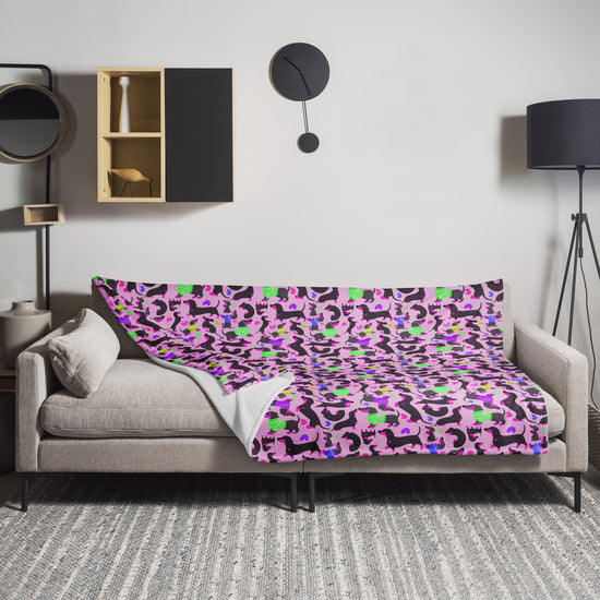 A modern living room features a beige sofa with a My Favourite Colour is Rainbow Silly Sausages Throw Blanket in pink, showcasing vibrant green and black patterns. The space also includes a round wall clock, minimalistic shelf decor, a black floor lamp, and a textured gray rug.