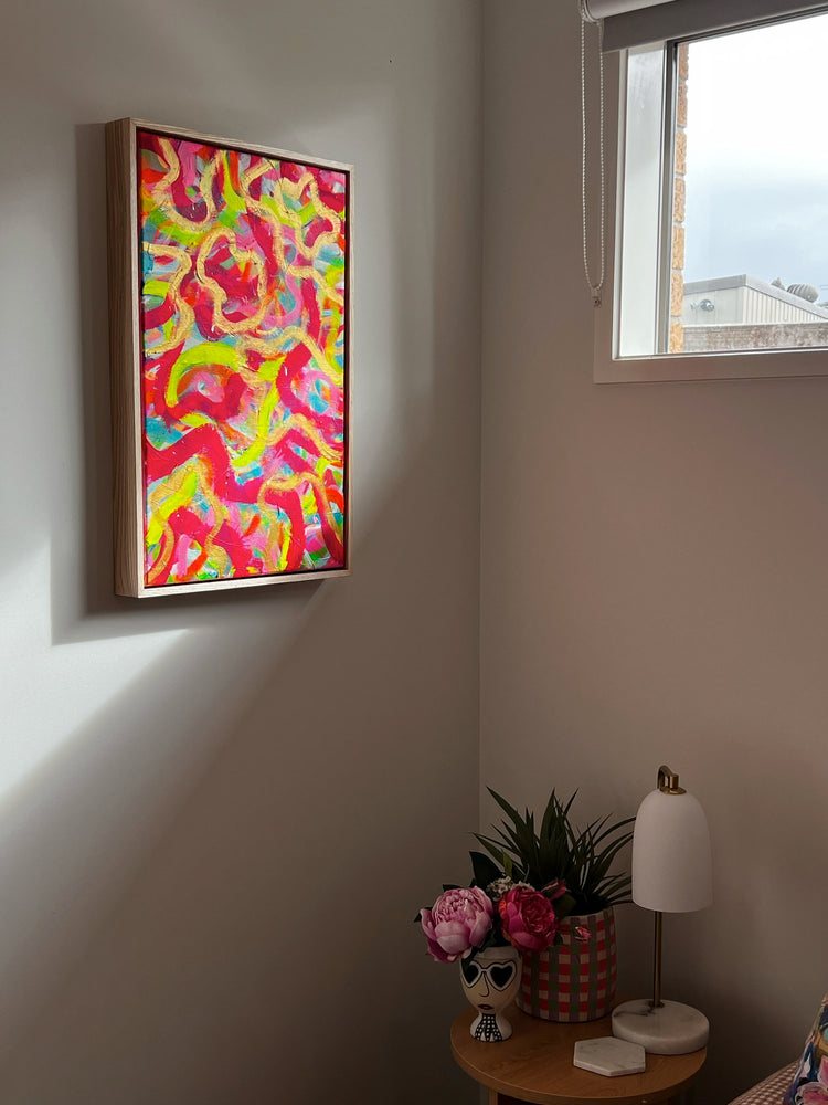 The Spaghetti Junction painting from My Store hangs on the white wall, lending texture and color. Below, a wooden table showcases a white lamp and decorative vase with pink flowers. A window above casts natural light across the room, enhancing its vibrant ambiance.