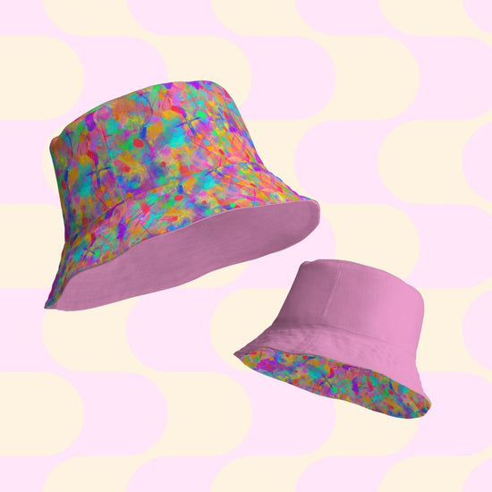 Two bucket hats on a pastel background: the Splotches and Crosses by My Favourite Colour is Rainbow, a reversible multicolored design in various sizes, and a solid pink hat. The backdrop of light yellow and pale pink shapes complements your style BFF perfectly.