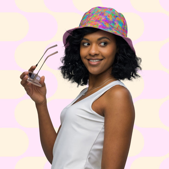 A woman with curly hair wears the Splotches and Crosses Reversible Bucket Hat by My Favourite Colour is Rainbow, paired with a white sleeveless top. Holding sunglasses, she smiles and gazes to the side against a retro pink and cream patterned background.