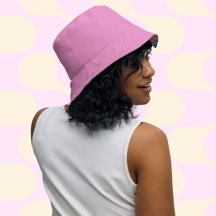 A curly-haired person wears the Splotches and Crosses Reversible Bucket Hat by My Favourite Colour is Rainbow and a white sleeveless top, looking over their shoulder against a pink and cream polka dot background.
