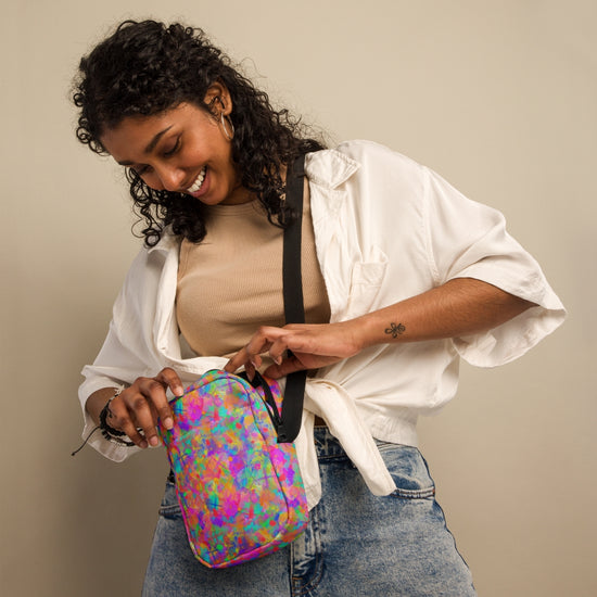 A smiling woman with curly hair, wearing a beige top, white shirt, and jeans, unzips the Splotches and Crosses Mini Crossbody Bag by My Favourite Colour is Rainbow. Shes standing against a neutral background with her gaze down.
