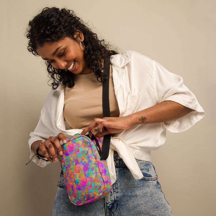 A smiling woman with curly hair, wearing a beige top, white shirt, and jeans, unzips the Splotches and Crosses Mini Crossbody Bag by My Favourite Colour is Rainbow. Shes standing against a neutral background with her gaze down.