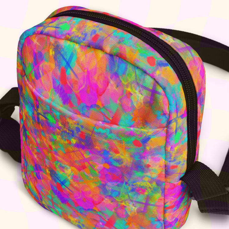 The Splotches and Crosses Mini Crossbody Bag by My Favourite Colour is Rainbow boasts a vibrant abstract pattern with splashes of pink, orange, blue, and green. It features roomy pockets, adjustable black straps, a black zipper, and is set against a pale yellow background.