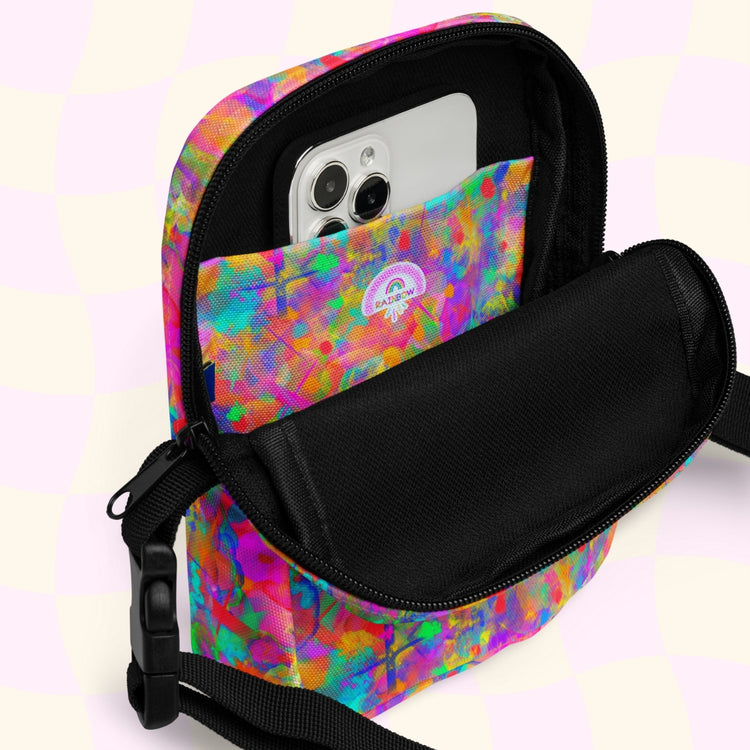 The open Splotches and Crosses Mini Crossbody Bag by My Favourite Colour is Rainbow reveals a smartphone inside. It features a vibrant abstract design, an adjustable black strap, and a zipper, against a soft checkered background.