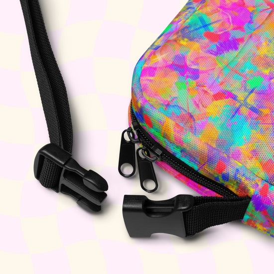 Close-up of the Splotches and Crosses Mini Crossbody Bag by My Favourite Colour is Rainbow, showcasing a vibrant multicolored abstract design with two black zippers and an adjustable strap with a plastic buckle, set against a light checkered background.