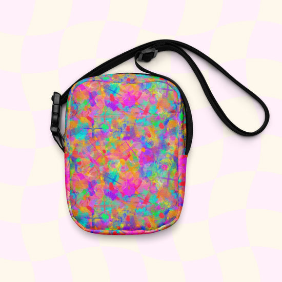 The Splotches and Crosses Mini Crossbody Bag by My Favourite Colour is Rainbow features a vibrant abstract design with splashes of pink, blue, green, and orange. It has thin black adjustable straps against a pastel checkered backdrop, making it perfect for any outing.