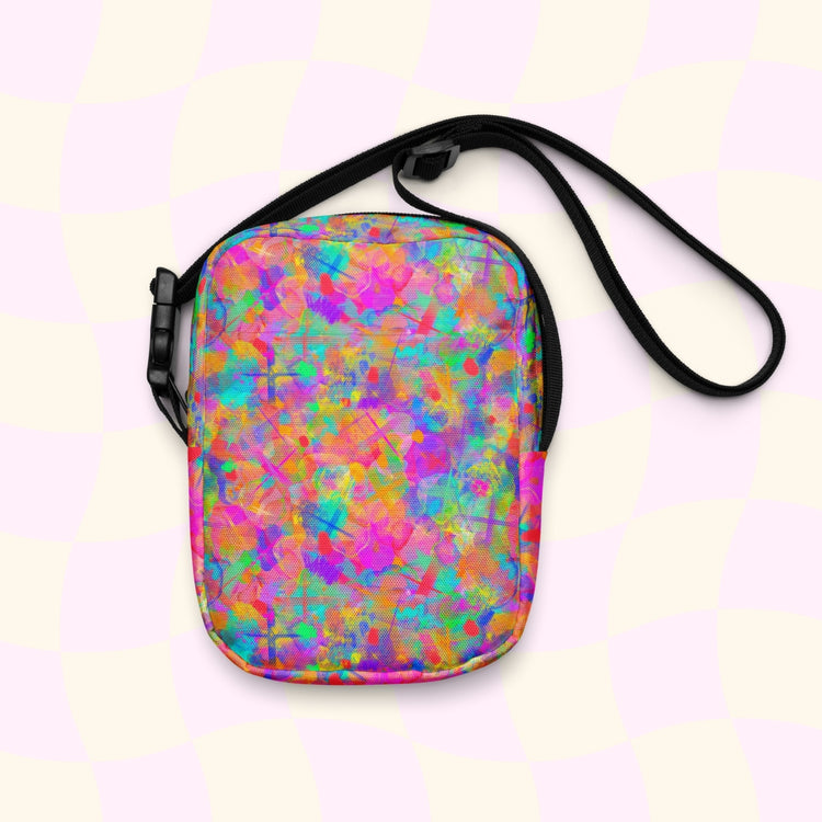 The Splotches and Crosses Mini Crossbody Bag by My Favourite Colour is Rainbow features a vibrant abstract design with splashes of pink, blue, green, and orange. It has thin black adjustable straps against a pastel checkered backdrop, making it perfect for any outing.