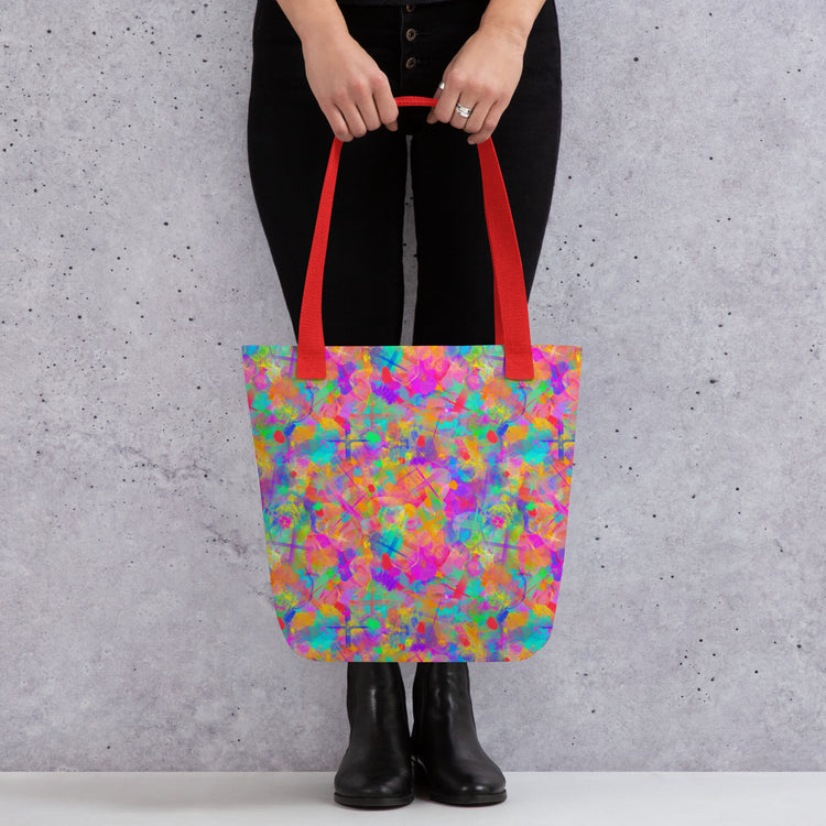 A person wearing black pants and boots carries a red-handled Splotches and Crosses Tote Bag by My Favourite Colour is Rainbow.  The bag features an abstract pattern of pink, blue, green, and orange splashes