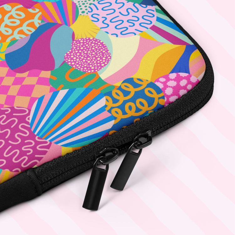 A close-up of the Squiggs and Stripes Laptop Sleeve by My Favourite Colour is Rainbow which features an abstract design of colourful geometric shapes, stripes and squiggles in pink, blue, orange yellow, and green. It includes a padded zipper binding with black dual pull tabs  for added convenience.  The print is on only one side of the laptop sleeve, the other side is black.