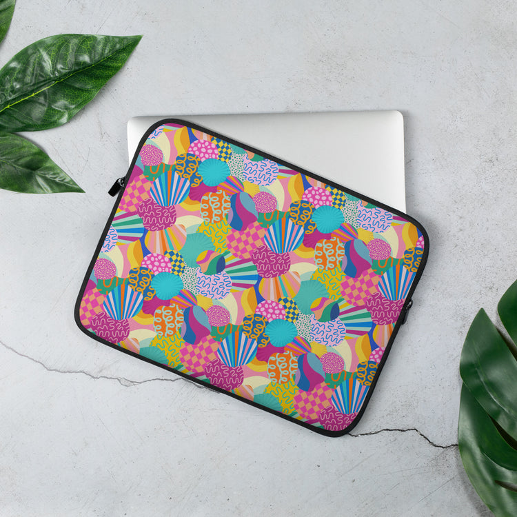 The 13 inch My Favourite Colour is Rainbow Squiggs and Stripes Laptop Sleeve features an abstract design of colourful geometric shapes, stripes and squiggles in pink, blue, orange yellow, and green. It rests on a light grey surface, partially covering a silver laptop.  There are scattered green leaves around.