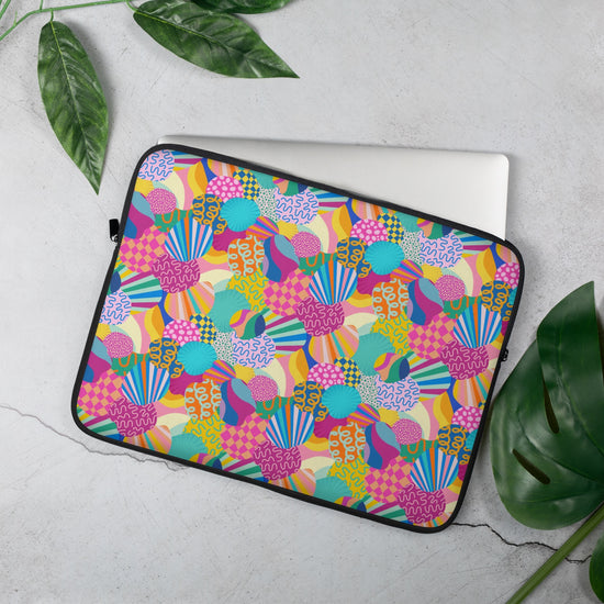 The 15 inch My Favourite Colour is Rainbow Squiggs and Stripes Laptop Sleeve features an abstract design of colourful geometric shapes, stripes and squiggles in pink, blue, orange yellow, and green. It rests on a light grey surface, partially covering a silver laptop.  There are scattered green leaves around.