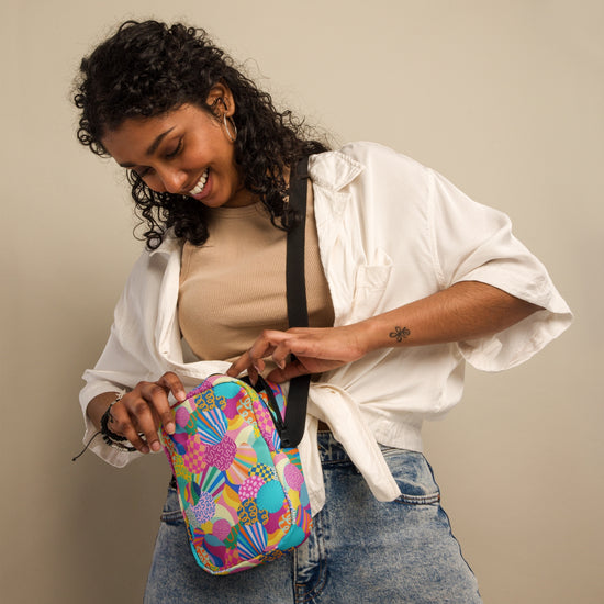 A woman with curly hair smiles while opening a Squiggs and Stripes Mini Crossbody bag by My Favourite Colour is Rainbow. The colorful, patterned bag has adjustable straps and she wears a white shirt over a beige top with jeans against a plain background.
