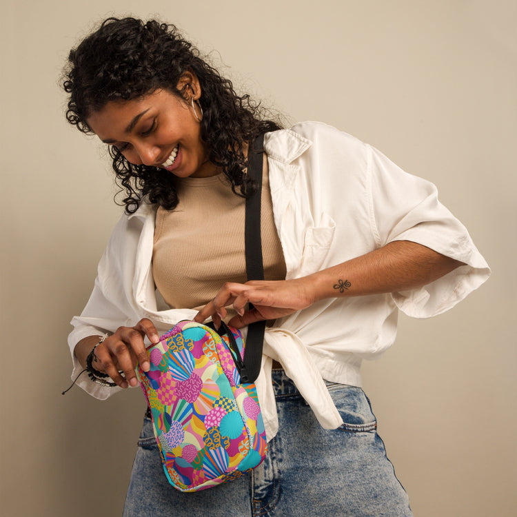 A woman with curly hair smiles while opening a Squiggs and Stripes Mini Crossbody bag by My Favourite Colour is Rainbow. The colorful, patterned bag has adjustable straps and she wears a white shirt over a beige top with jeans against a plain background.