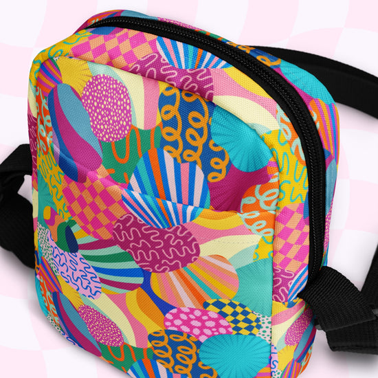 The Squiggs and Stripes Mini Crossbody bag by My Favourite Colour is Rainbow showcases vibrant geometric shapes, wavy lines, and circular designs in pink, blue, yellow, and green. It features black adjustable straps and a zippered closure on a checkered background.