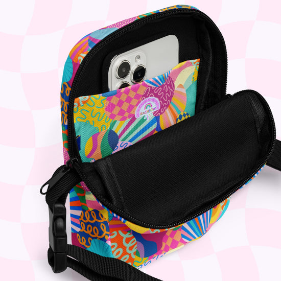 The Squiggs and Stripes Mini Crossbody bag by My Favourite Colour is Rainbow features a colorful design with abstract patterns, adjustable straps, and a front pocket for holding a smartphone. Its open black zipper reveals the phone and bag tag against a charming light pink checkered background.