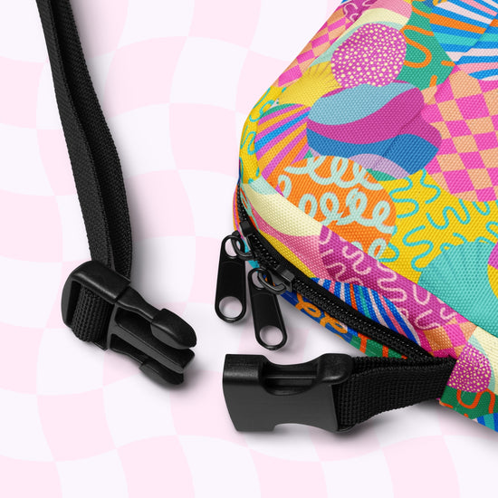 Close-up of a Squiggs and Stripes Mini Crossbody bag by My Favourite Colour is Rainbow. The colorful, water-resistant bag features abstract pink, blue, and yellow patterns with black zippers and adjustable straps with a plastic buckle against a light pink checkered background.