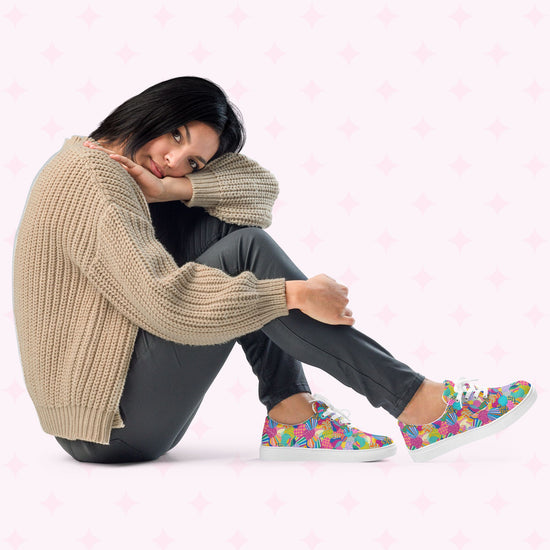A woman sits cross-legged on the floor wearing a beige knitted sweater, dark pants, and Squiggs and Stripes Canvas Sneakers by My Favourite Colour is Rainbow. Her head rests on her knees against a light background with subtle star patterns, showcasing her vibrant style.