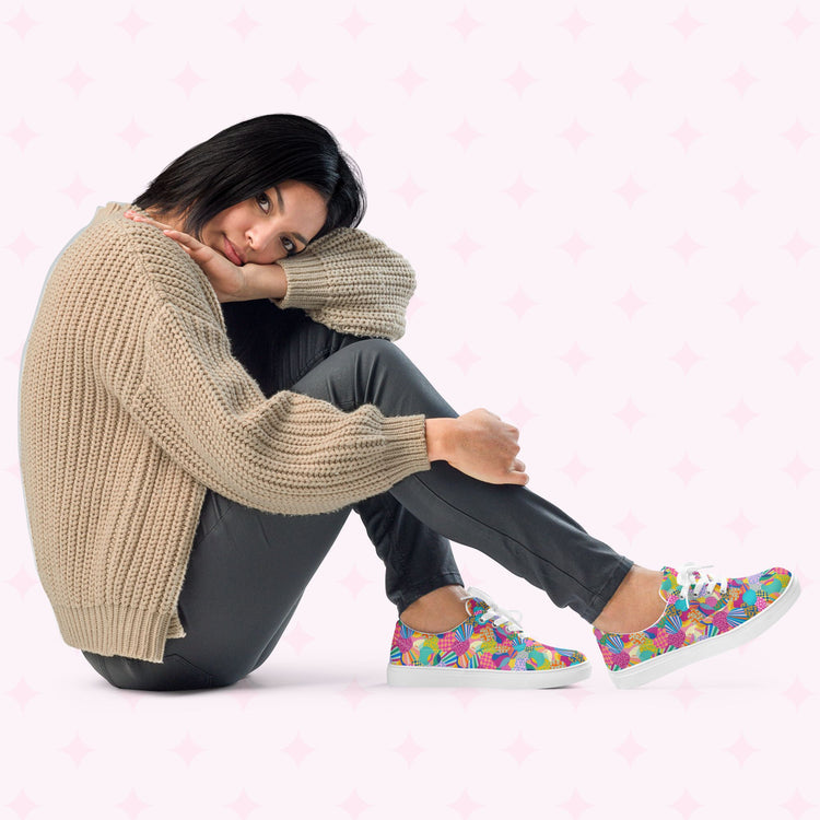 A woman sits cross-legged on the floor wearing a beige knitted sweater, dark pants, and Squiggs and Stripes Canvas Sneakers by My Favourite Colour is Rainbow. Her head rests on her knees against a light background with subtle star patterns, showcasing her vibrant style.