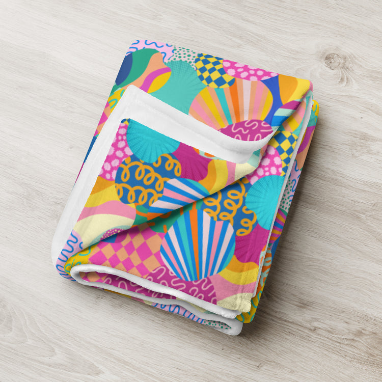 The Squiggs and Stripes Throw Blanket by My Favourite Colour is Rainbow, featuring a vibrant abstract design with stripes and swirls in pink, yellow, blue, and green, rests on a light wood surface.
