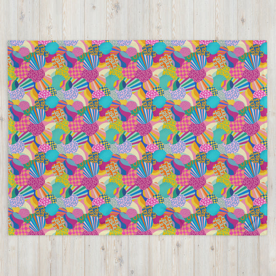 The Squiggs and Stripes Throw Blanket by My Favourite Colour is Rainbow features a vibrant design of overlapping geometric shapes, stripes, and polka dots in pink, blue, yellow, and green on a light wooden background.